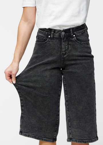 Model strækker de løse, mørkegrå denim capris i farven Shadows ved hoften for at vise materialets stretch.
