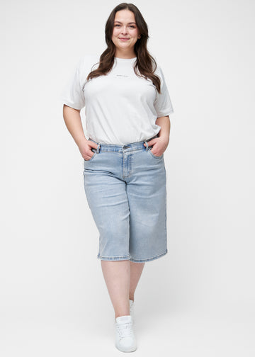 Helbild av plus-size modell i lösa, ljusblå denim capris i färgen Waves som går till precis under knät.