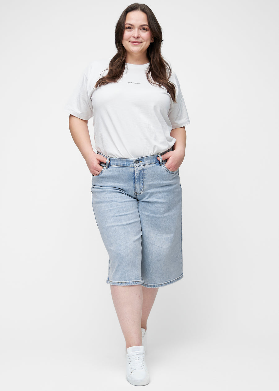 Helbild av plus-size modell i lösa, ljusblå denim capris i färgen Waves som går till precis under knät.