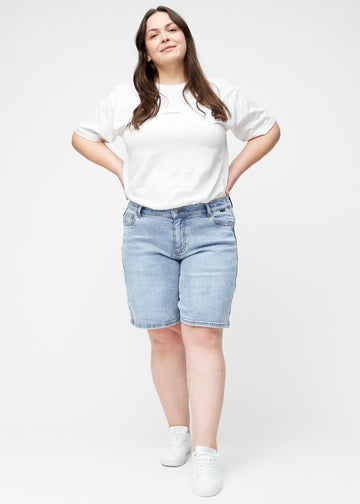 Helbild av plus-size modell i lösa, ljusblå denimshorts i färgen Waves som går till precis över knät.