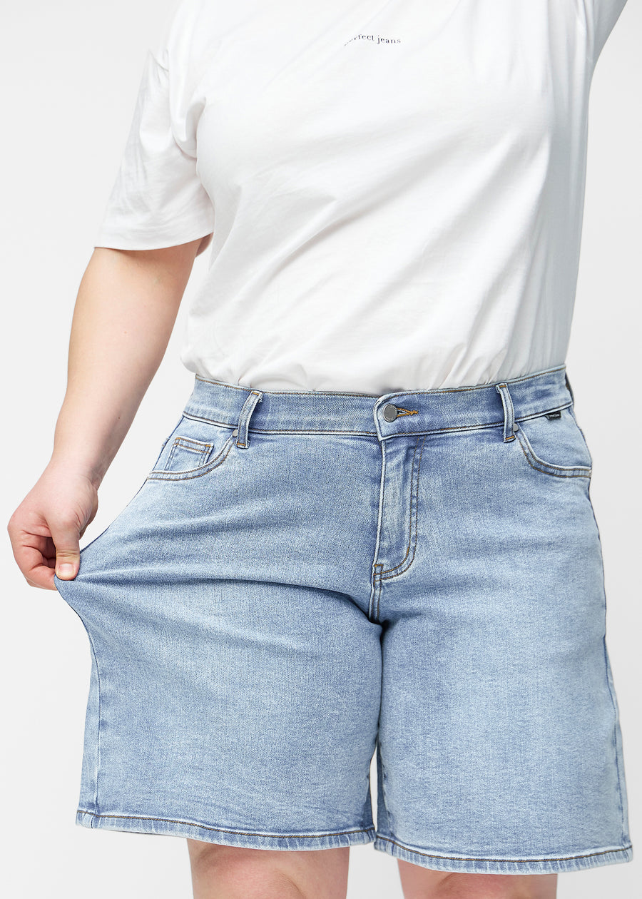 Plus-size modell stretchar de lösa, ljusblå denimshortsen i färgen Waves vid höften för att visa materialets stretch.