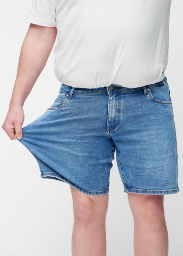 Plus-size modell sträcker blå denimshorts för herr i färgen Rivers vid låret för att visa materialets stretch.