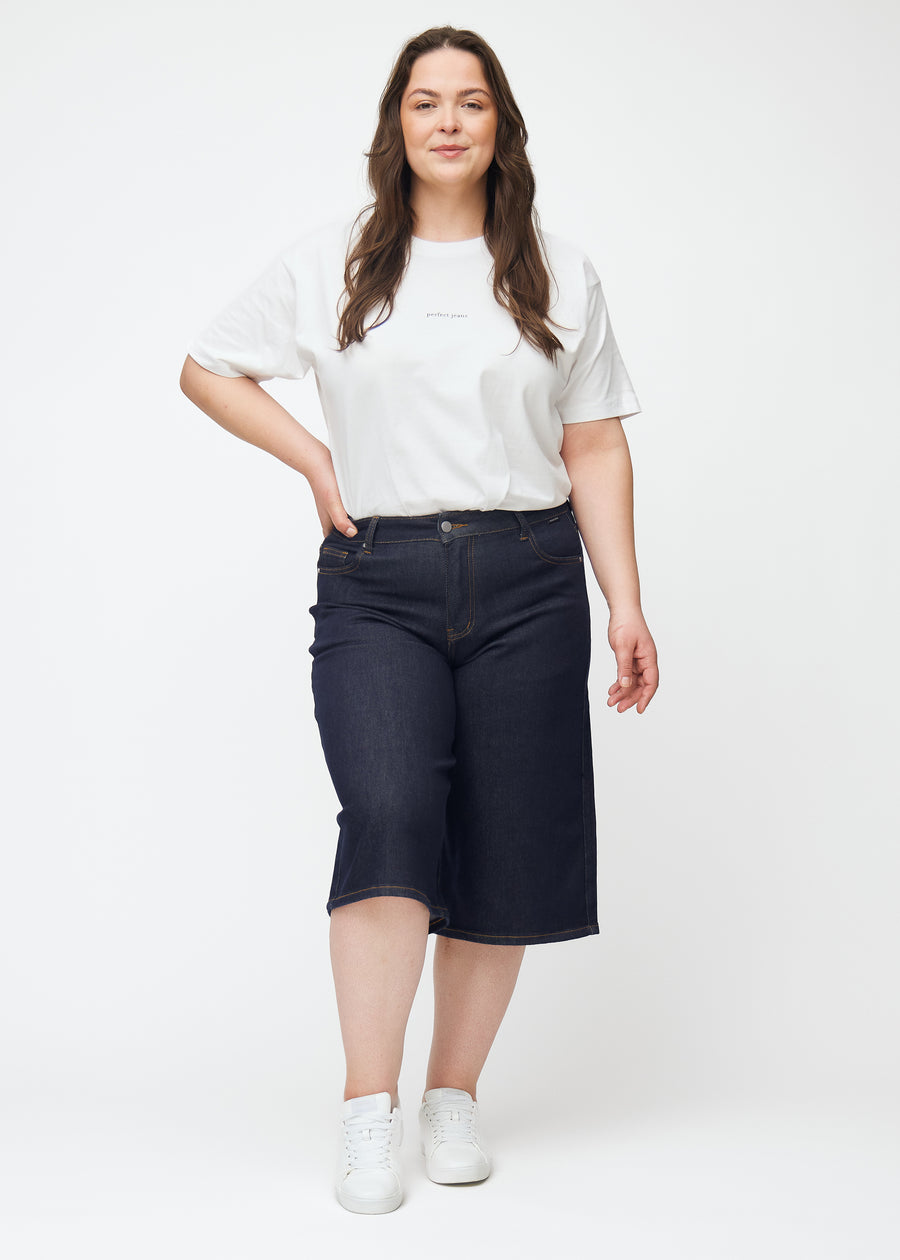 Helbild av plus-size modell i mörkblå denim capris för dam i färgen Midnights med loose passform.