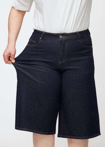 Plus-size modell sträcker mörkblå denim capris för dam i färgen Midnights vid låret för att visa materialets stretch.