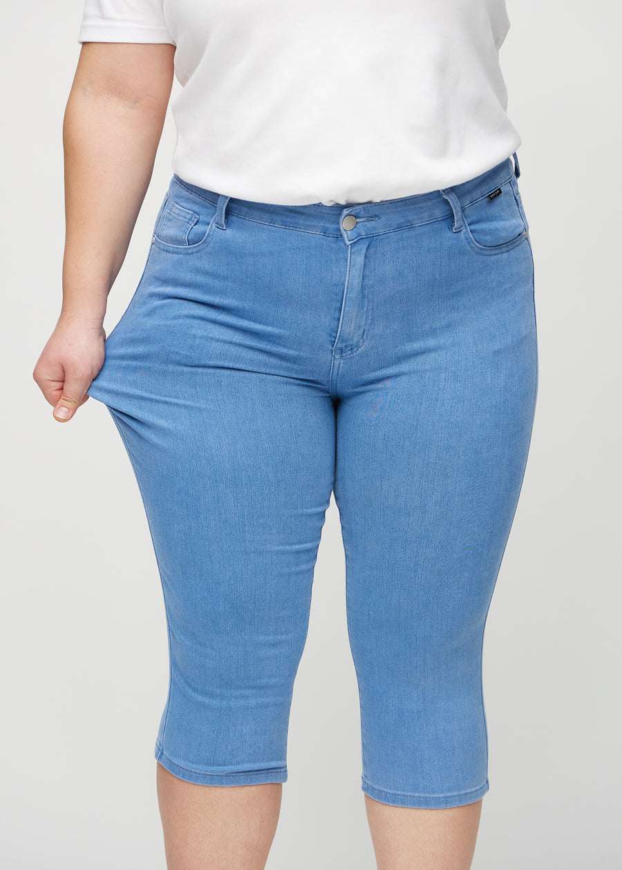 Skinnycapris för kvinnor i blått jeanstyg, färgen Geraniums. En plus size-modell drar i tyget för att visa flexibiliteten.