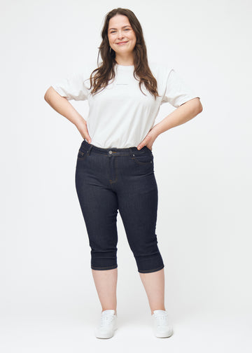 Helbild av plus-size modell i mörkblå denim capris för dam i färgen Midnights med skinny passform.