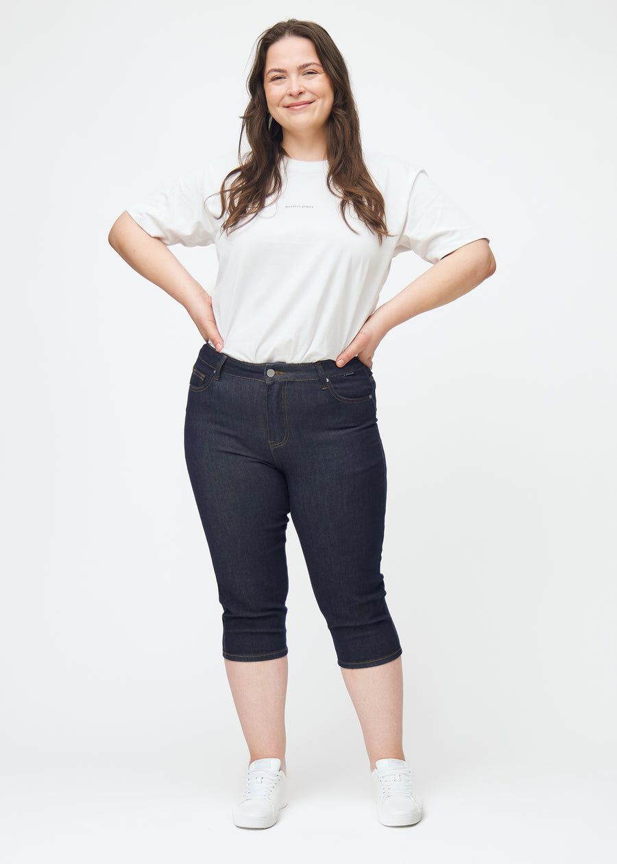 Helbild av plus-size modell i mörkblå denim capris för dam i färgen Midnights med skinny passform.