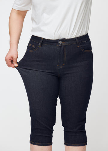 Plus-size modell sträcker mörkblå denim capris för dam i färgen Midnights vid låret för att visa stretch.