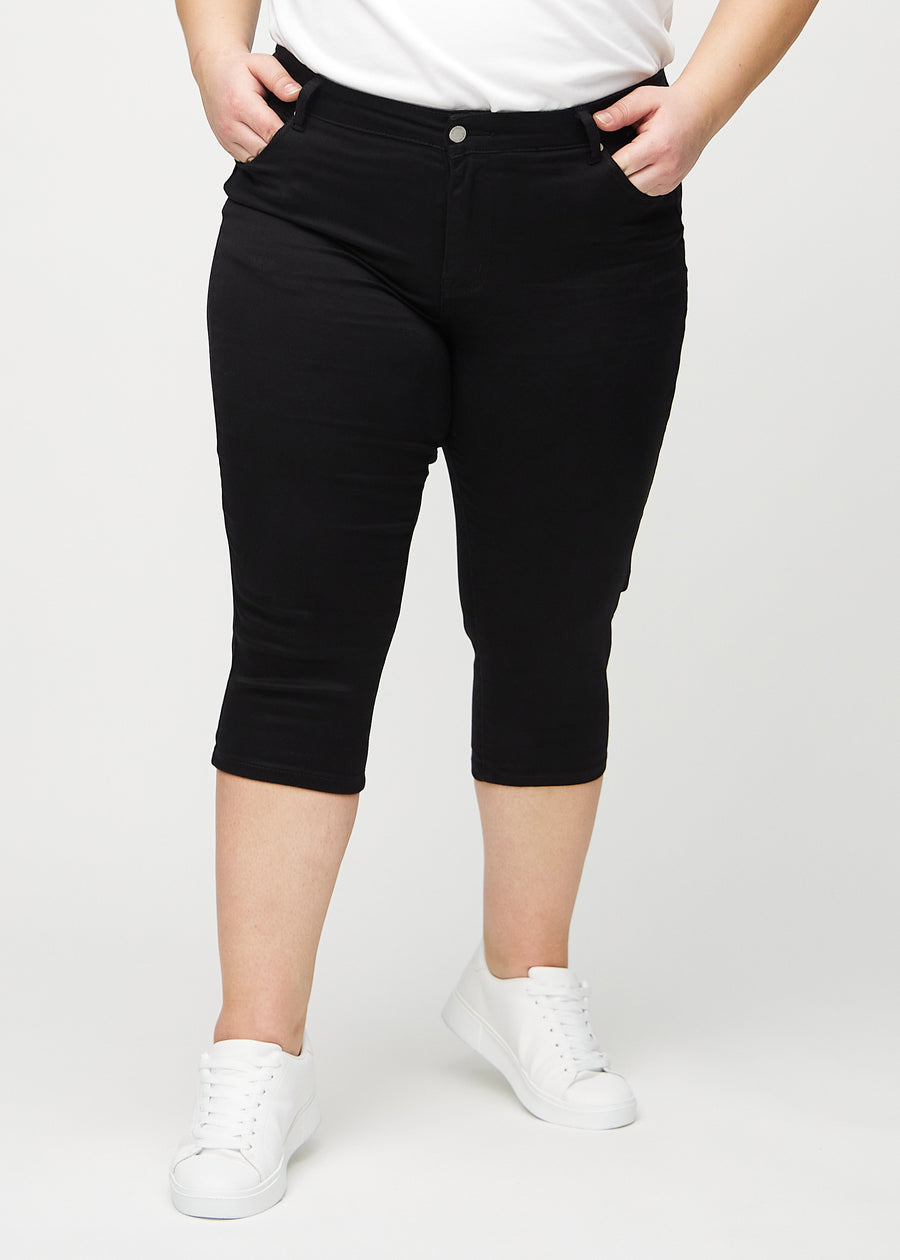 Svarta skinny capris, modellnamn Ravens, som sitter tätt längs benet och slutar precis under knät på en plus-size-modell, framifrån.