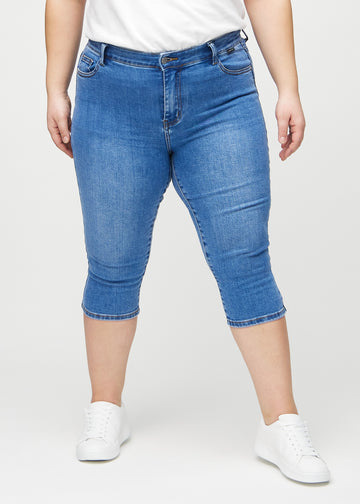 Denim skinny capris, modellnamn Rivers, som sitter tätt längs benet och slutar precis under knät på en plus size-modell, sedd framifrån.