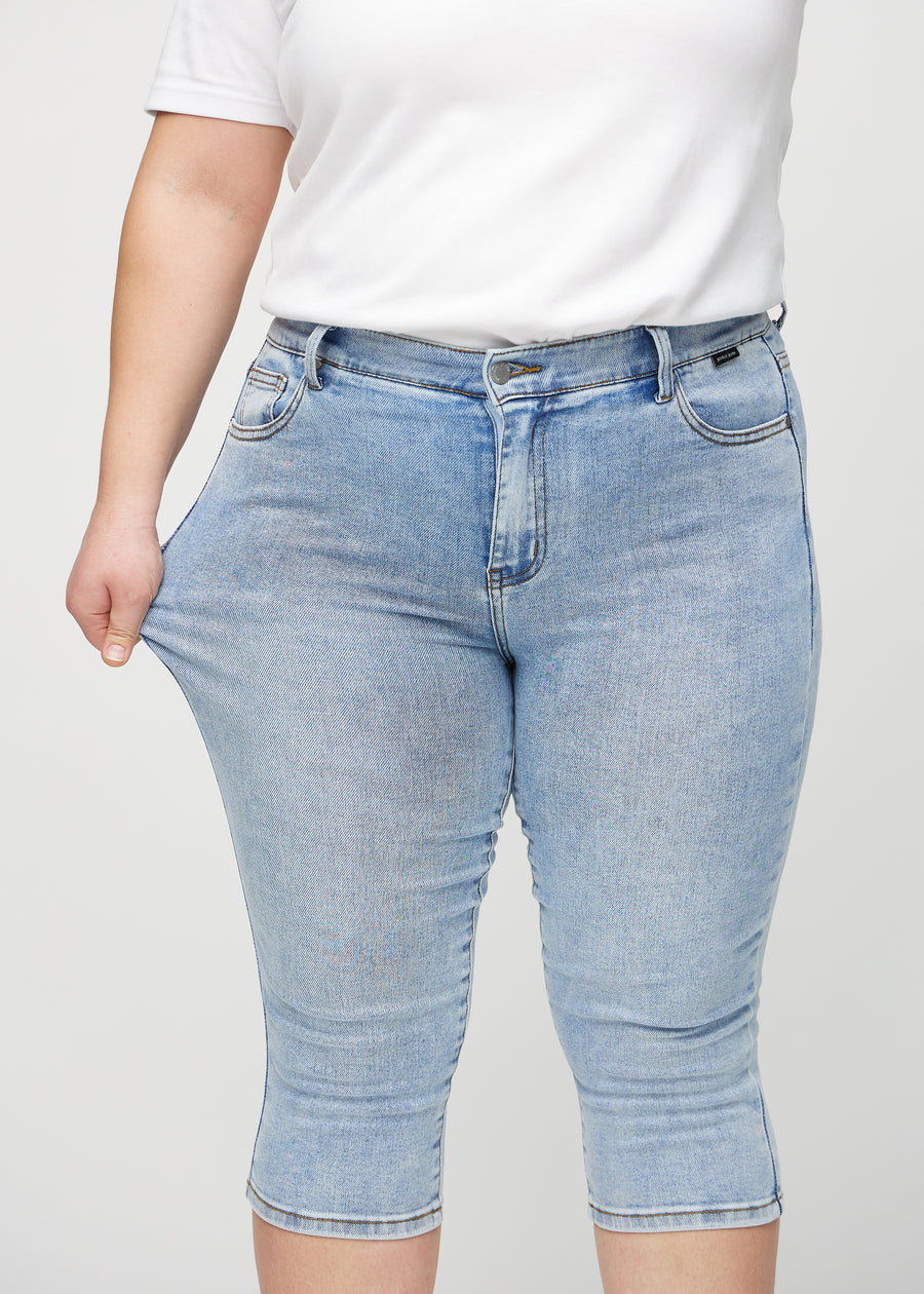 Skinnycapris för kvinnor i ljusblå denim, färgen Waves. En plus size-modell drar i tyget för att visa flexibiliteten.