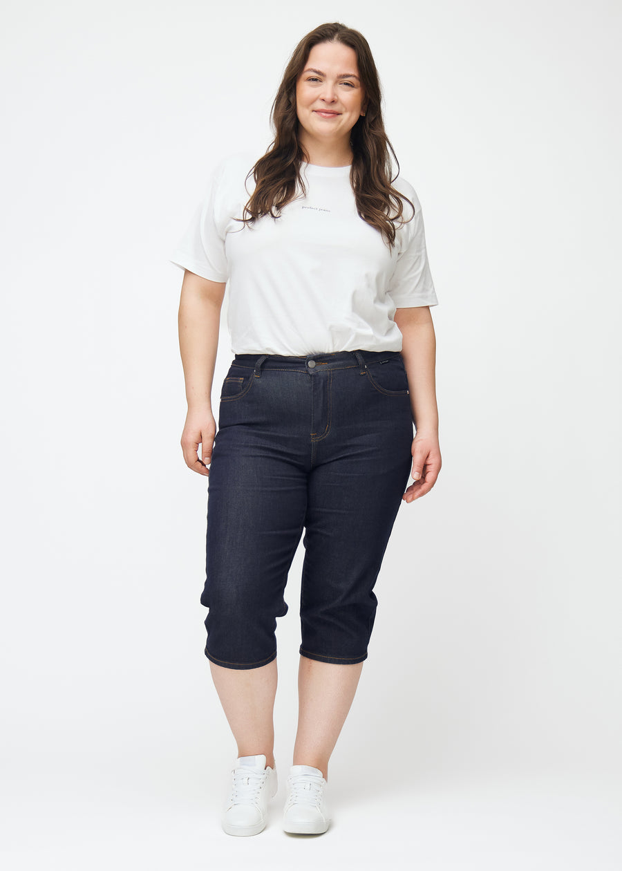 Helbild av plus-size modell i mörkblå denim capris för dam i färgen Midnights med slim passform.