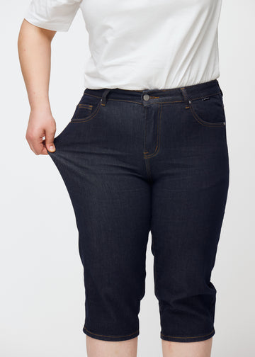 Plus-size modell sträcker mörkblå denim capris för dam i färgen Midnights vid låret för att visa stretch.