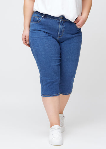 Slimcapris för kvinnor i mörkblå denim, färgen Oceans. Framifrån på en plus size-modell, med figursydd passform till strax under knät.