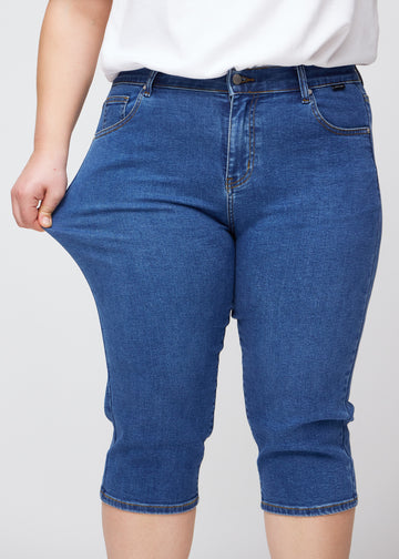 Slimcapris för kvinnor i mörkblå denim, färgen Oceans. En plus size-modell drar i tyget vid låret för att visa stretch och flexibilitet.