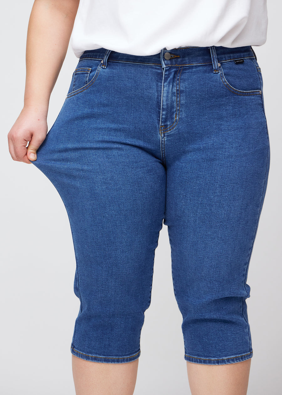 Slimcapris för kvinnor i mörkblå denim, färgen Oceans. En plus size-modell drar i tyget vid låret för att visa stretch och flexibilitet.