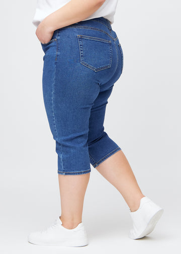 Slimcapris för kvinnor i mörkblå denim, färgen Oceans. Från sidan på en plus size-modell, med figursydd passform och medelhög midja (Mid Rise).
