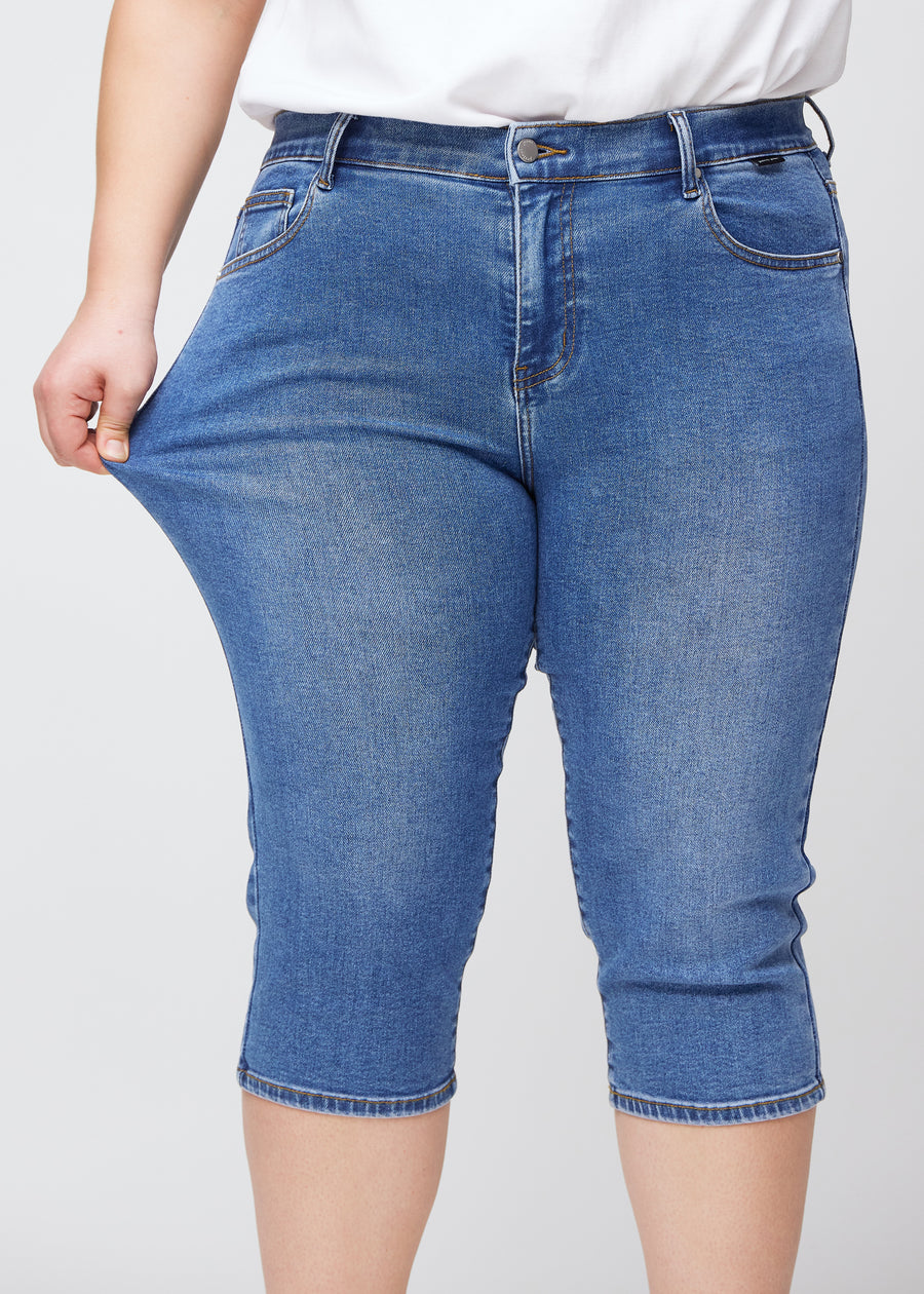 Slimcapris för kvinnor i blå denim, färgen Rivers. En plus size-modell drar i tyget vid låret för att visa stretch och flexibilitet.