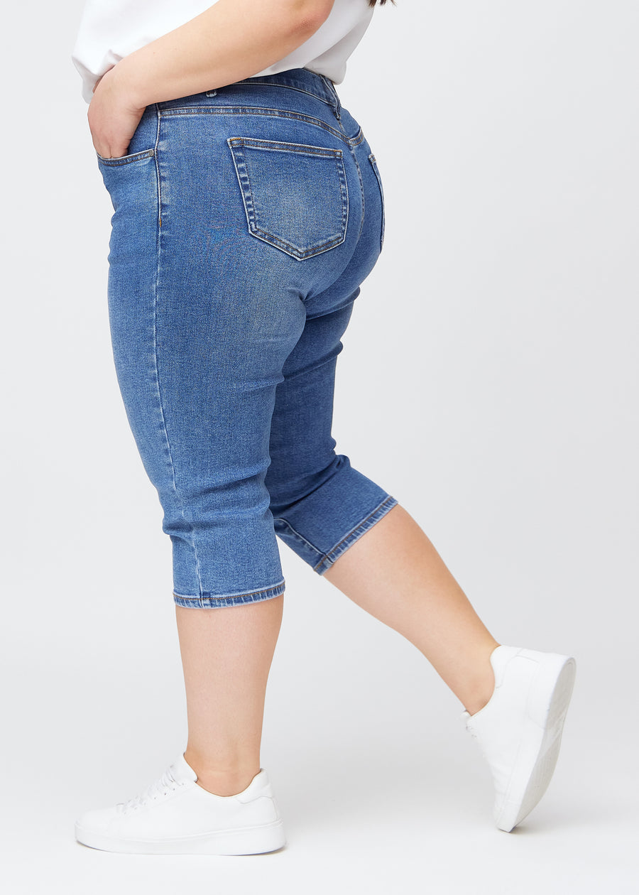 Slimcapris för kvinnor i blå denim, färgen Rivers. Från sidan på en plus size-modell, med figursydd passform och medelhög midja (Mid Rise).
