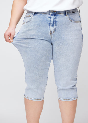 Slimcapris för kvinnor i ljusblå denim, färgen Waves. En plus size-modell drar i tyget vid låret för att visa stretch och flexibilitet.
