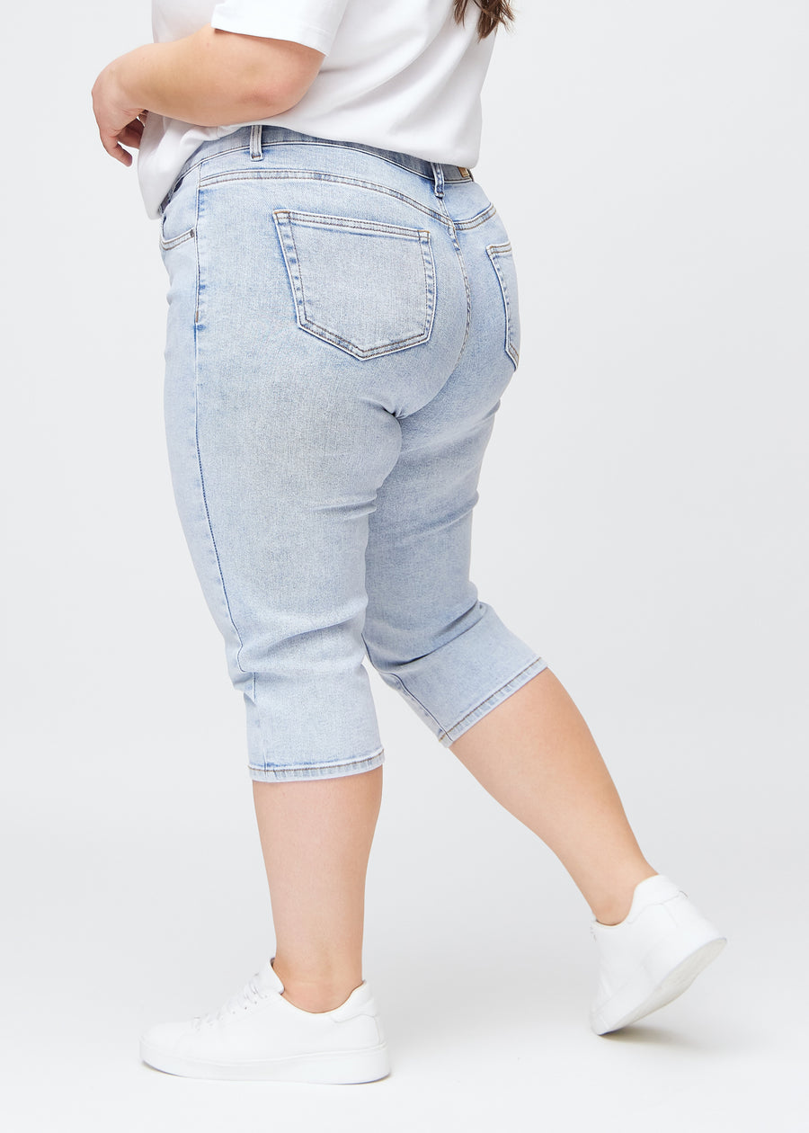 Slimcapris för kvinnor i ljusblå denim, färgen Waves. Från sidan på en plus size-modell, med figursydd passform och medelhög midja (Mid Rise).