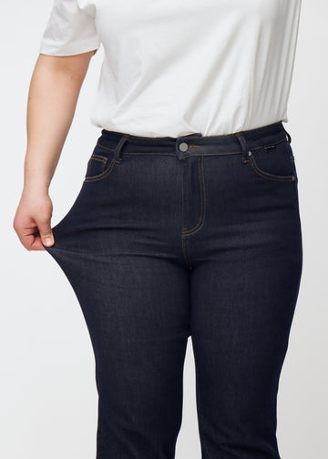 Plus size-modell sträcker djupt mörkblå denim jeans för kvinnor i färgen Midnights vid låret för att visa materialets stretch.