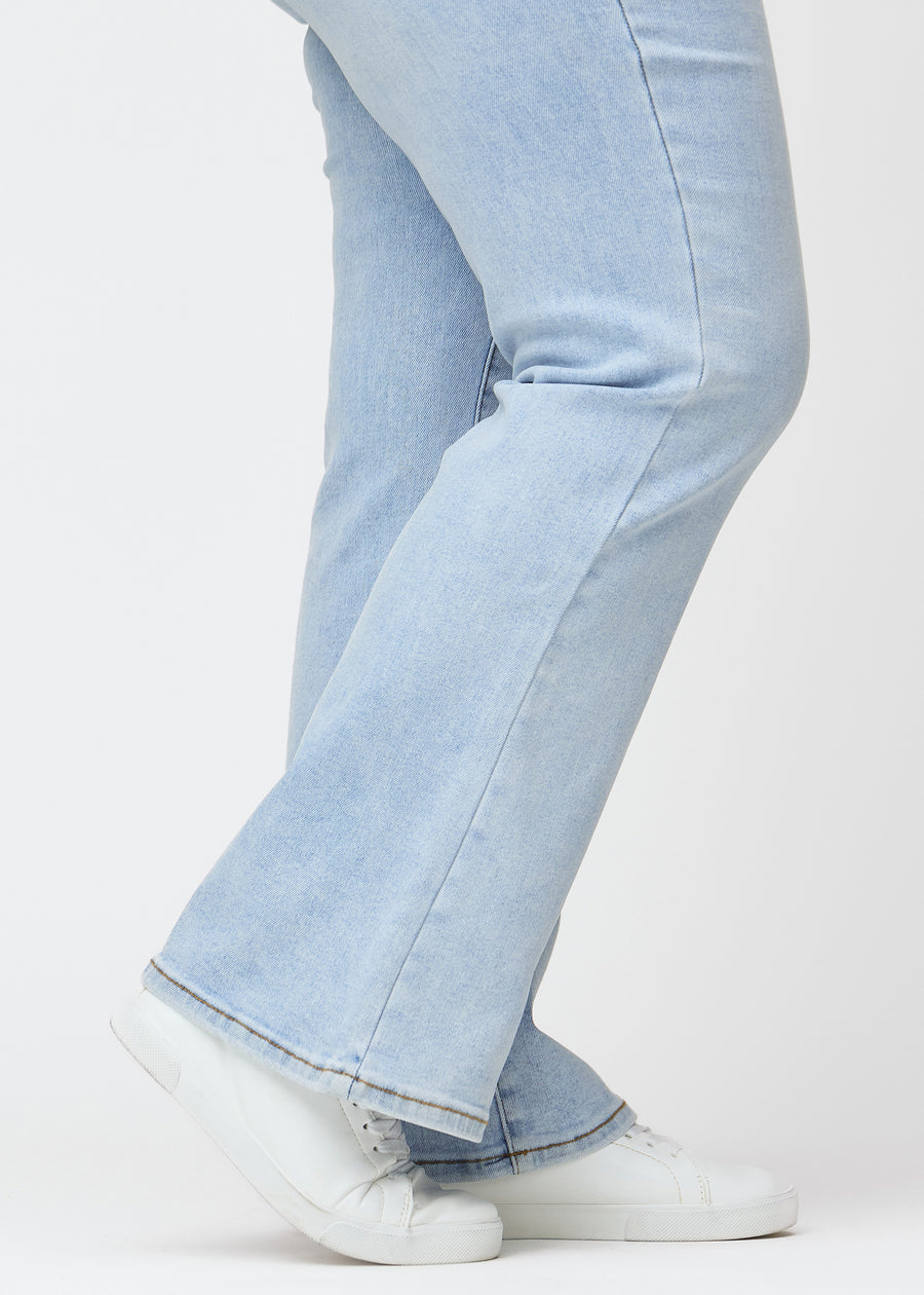 Ljusblå mid-rise bootcutjeans i färgen Waves. Närbild vid ankeln på en plus size-modell som visar den lätta utsvängningen nedtill.