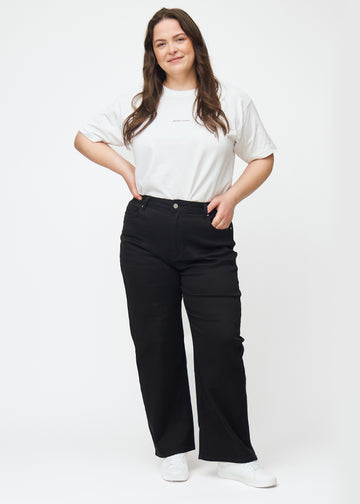 En helbild av en plus-size-modell i svarta högmidjade jeans, som sitter åt i midjan och har en lös passform med vidd nedtill.