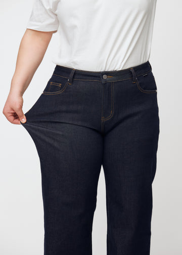 Plus-size-modell sträcker den djupt mörkblå denim jeans för kvinnor i färgen Midnights vid låret för att visa materialets stretch.