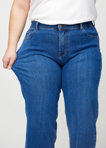 Loose jeans för kvinnor i mörkblå, färgen Oceans. En plus size-modell drar i jeanstyget vid låret för att visa stretchen.