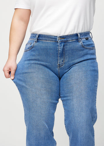 Loose jeans för kvinnor i blå, färgen Rivers. En plus size-modell drar i jeanstyget vid låret för att visa stretchen.