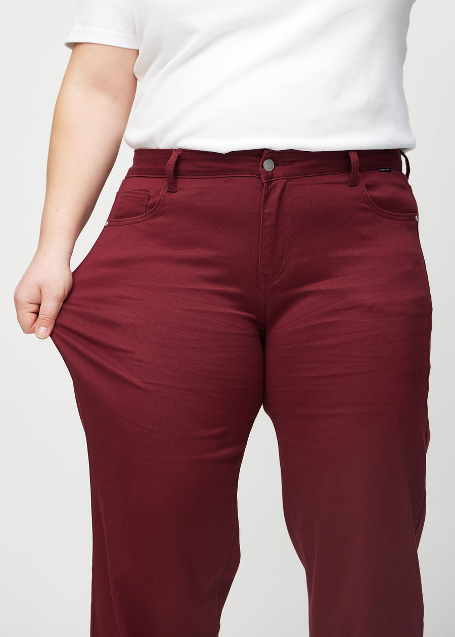 Loose jeans för kvinnor i mörkröd, färgen Santas. En plus size-modell drar i jeanstyget vid låret för att visa stretchen.