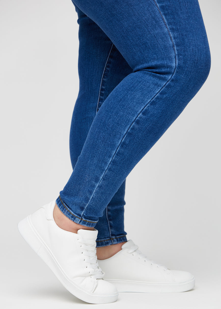 Skinnyjeans för kvinnor i mörkblå, färgen Oceans. Närbild vid anklarna på en plus size-modell som visar de åtsittande bensluten.