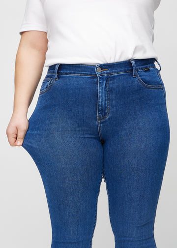 Skinnyjeans för kvinnor i mörkblå, färgen Oceans. En plus size-modell drar i jeanstyget vid låret för att visa stretchen.
