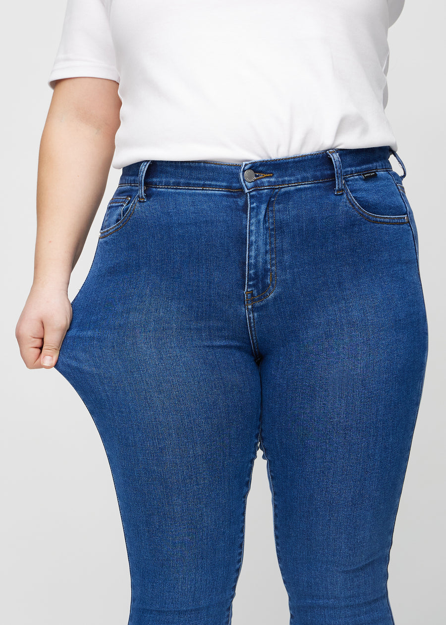 Skinnyjeans för kvinnor i mörkblå, färgen Oceans. En plus size-modell drar i jeanstyget vid låret för att visa stretchen.