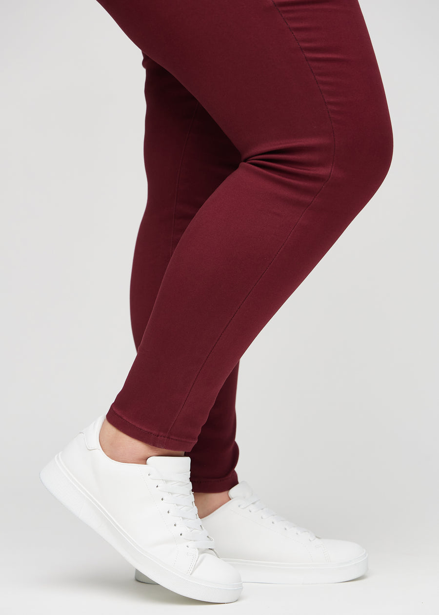 Skinnyjeans för kvinnor i mörkröd, färgen Santas. Närbild vid anklarna på en plus size-modell som visar de åtsittande bensluten.