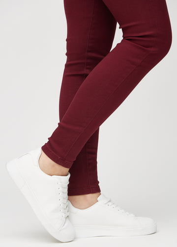 Skinnyjeans för kvinnor i mörkröd, färgen Santas. Närbild vid anklarna på en modell som visar de åtsittande bensluten.