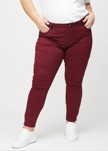 Skinnyjeans för kvinnor i mörkröd, färgen Santas. Framifrån på en plus size-modell, med nära passform runt benen.