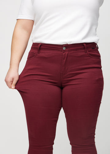 Skinnyjeans för kvinnor i mörkröd, färgen Santas. En plus size-modell drar i jeanstyget vid låret för att visa stretchen.