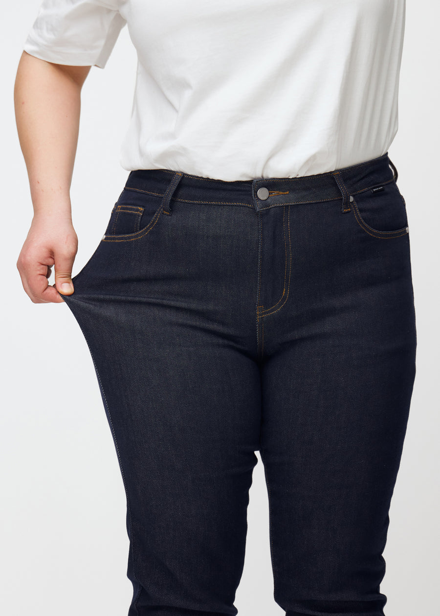 Plus-size-modell stretchar den djupt mörkblå denimen jeans för kvinnor i färgen Midnights vid låret för att visa materialets stretch.