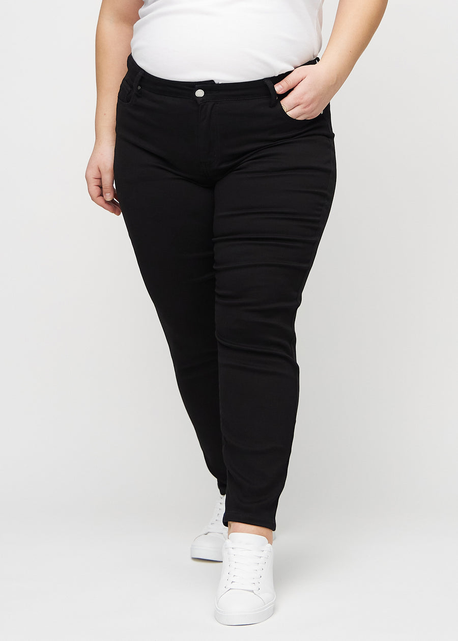 Slimjeans för kvinnor i svart, färgen Ravens. Framifrån på en plus size-modell, med kroppsnära ben och lite mer utrymme vid anklarna.