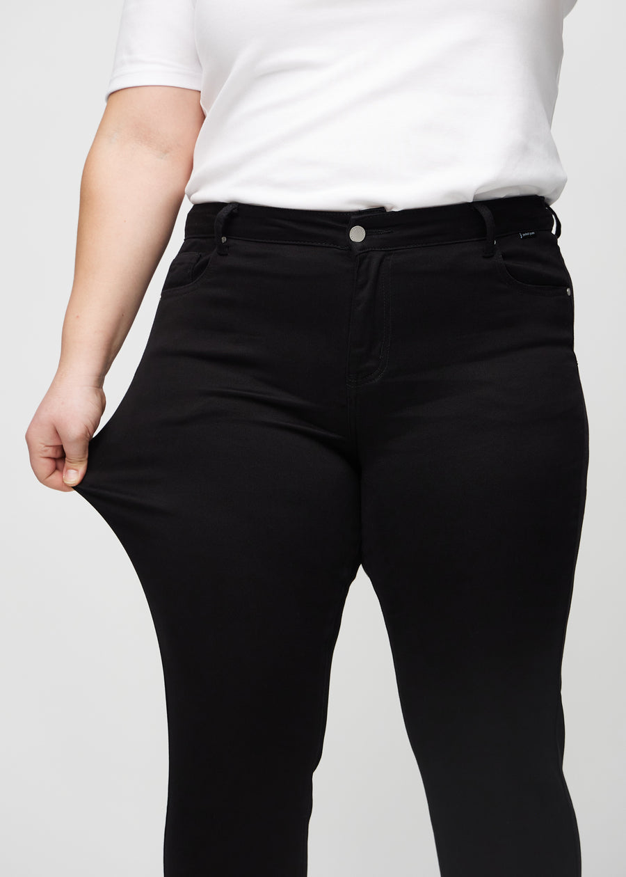 Slimjeans för kvinnor i svart, färgen Ravens. En plus size-modell drar i jeanstyget vid låret för att visa stretchen.