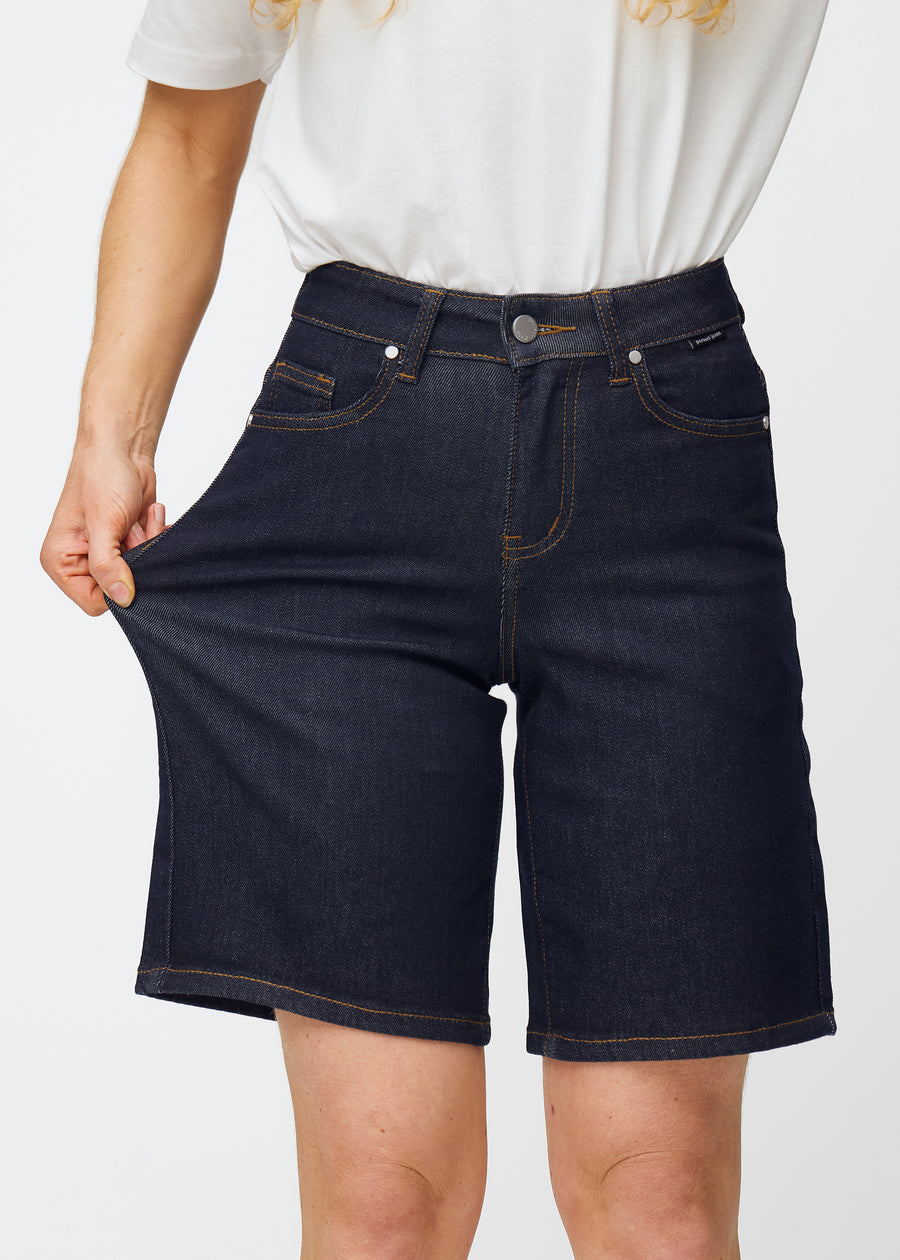 Modell sträcker mörkblå denimshorts för dam i färgen Midnights vid låret för att visa materialets stretch.