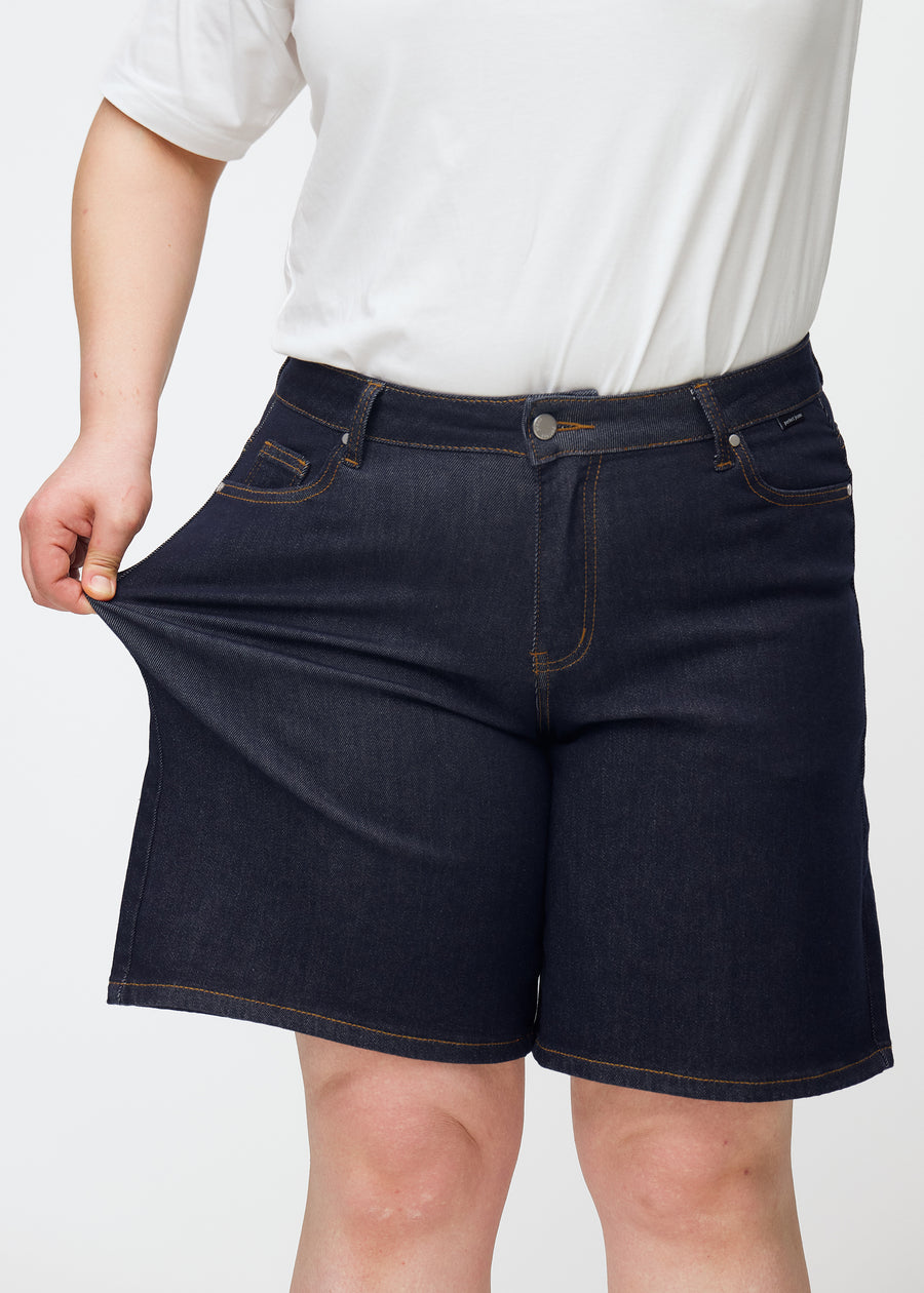 Plus-size modell sträcker mörkblå denimshorts för dam i färgen Midnights vid låret för att visa stretch.