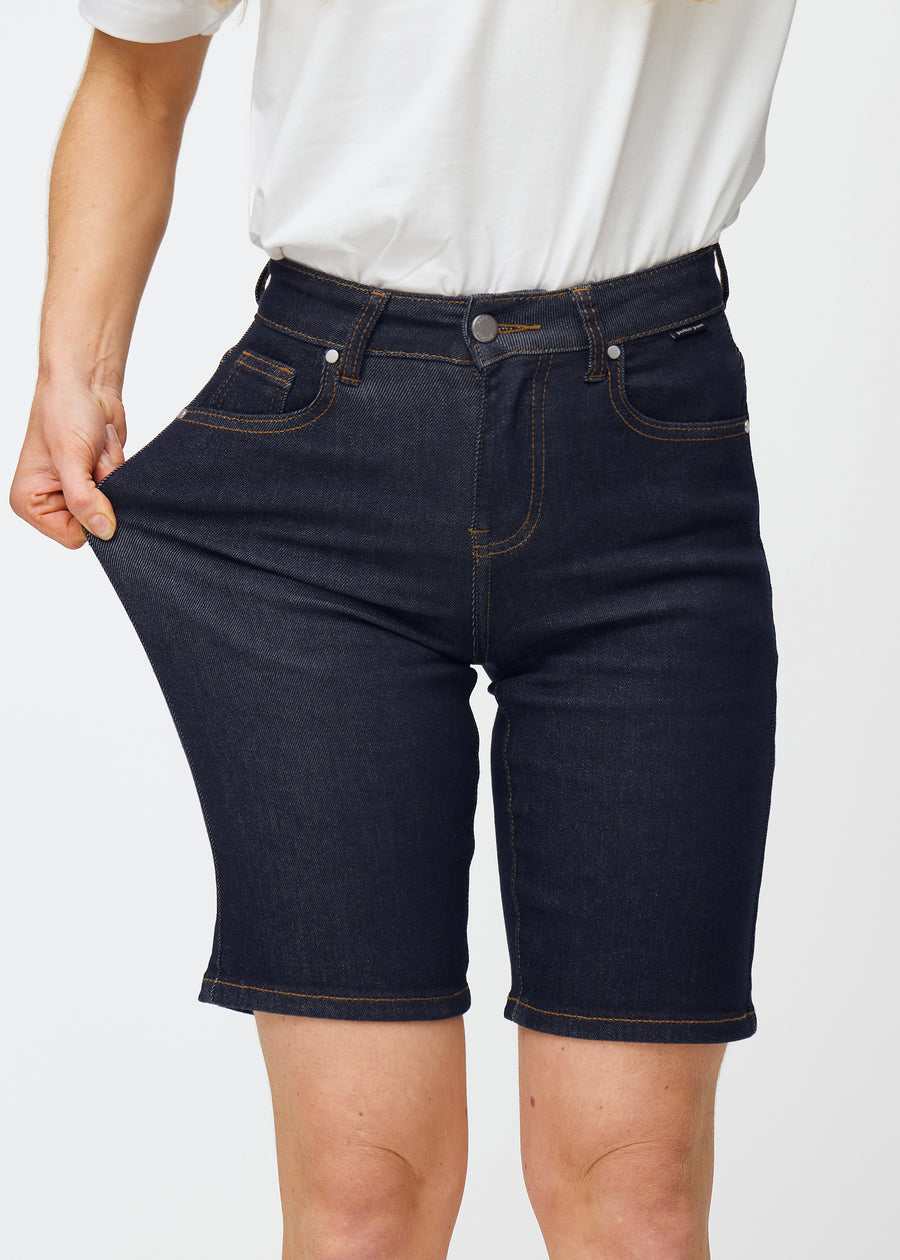 Modell sträcker mörkblå denimshorts för dam i färgen Midnights vid låret för att visa materialets stretch.