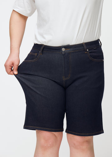 Plus-size modell sträcker mörkblå denimshorts för dam i färgen Midnights vid låret för att visa stretch.