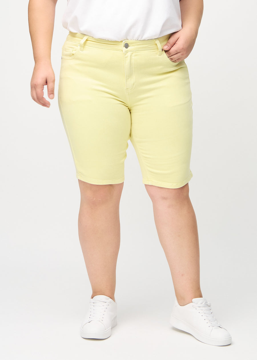 Ljusgula skinny middle shorts, modellnamn Lemonades, som sitter tätt över låret och slutar precis ovanför knät på en plus size-modell, sedda framifrån.