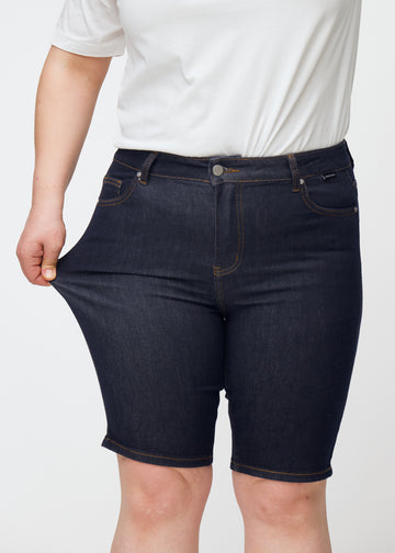 Plus-size modell sträcker mörkblå denimshorts för dam i färgen Midnights vid låret för att visa stretch.