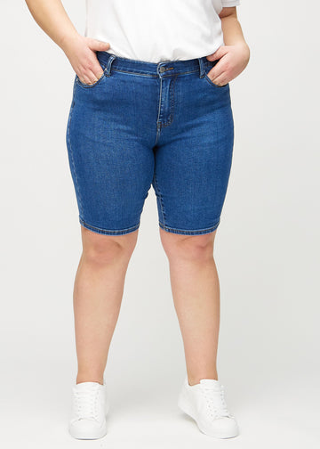 Mörk denim skinny middle shorts, modellnamn Oceans, som sitter tätt över låret och slutar precis ovanför knät på en plus-size-modell, framifrån.