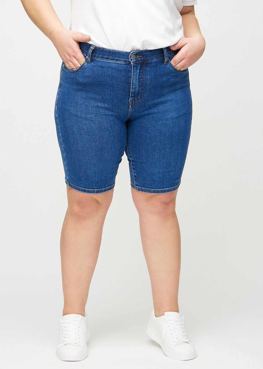 Mörk denim skinny middle shorts, modellnamn Oceans, som sitter tätt över låret och slutar precis ovanför knät på en plus-size-modell, framifrån.